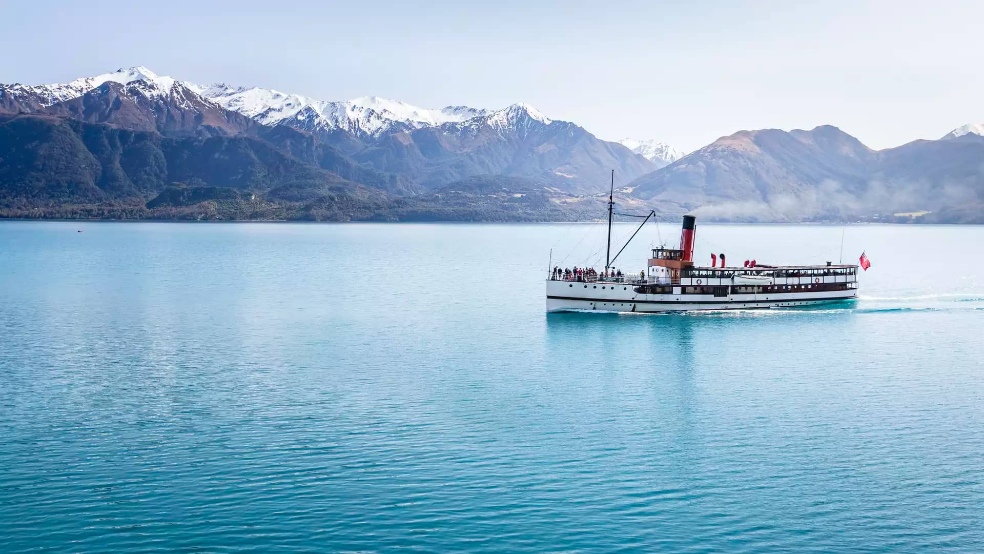 Steamboat cruising on blue lake water with snowy mountains in the background on a clear day. Credit: Real Journeys