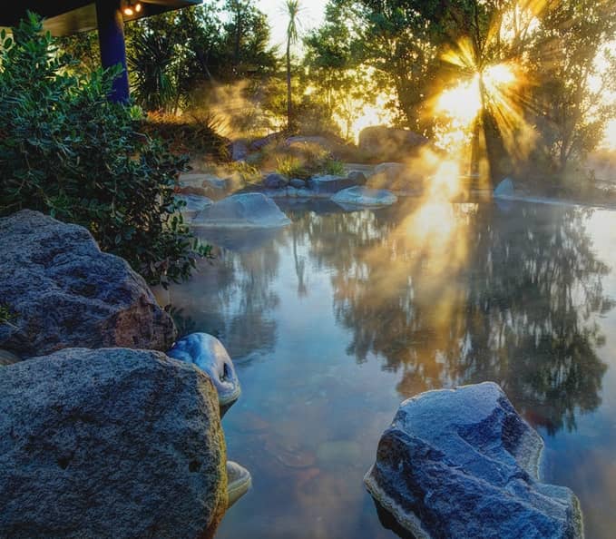 Outdoor hot spring spa surrounded by rocks and trees with steam rising and sunlight shining through.