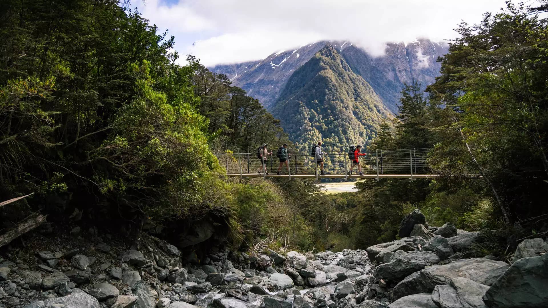 Four hikers crossing a suspension bridge over rocky riverbed with mountains and forest behind