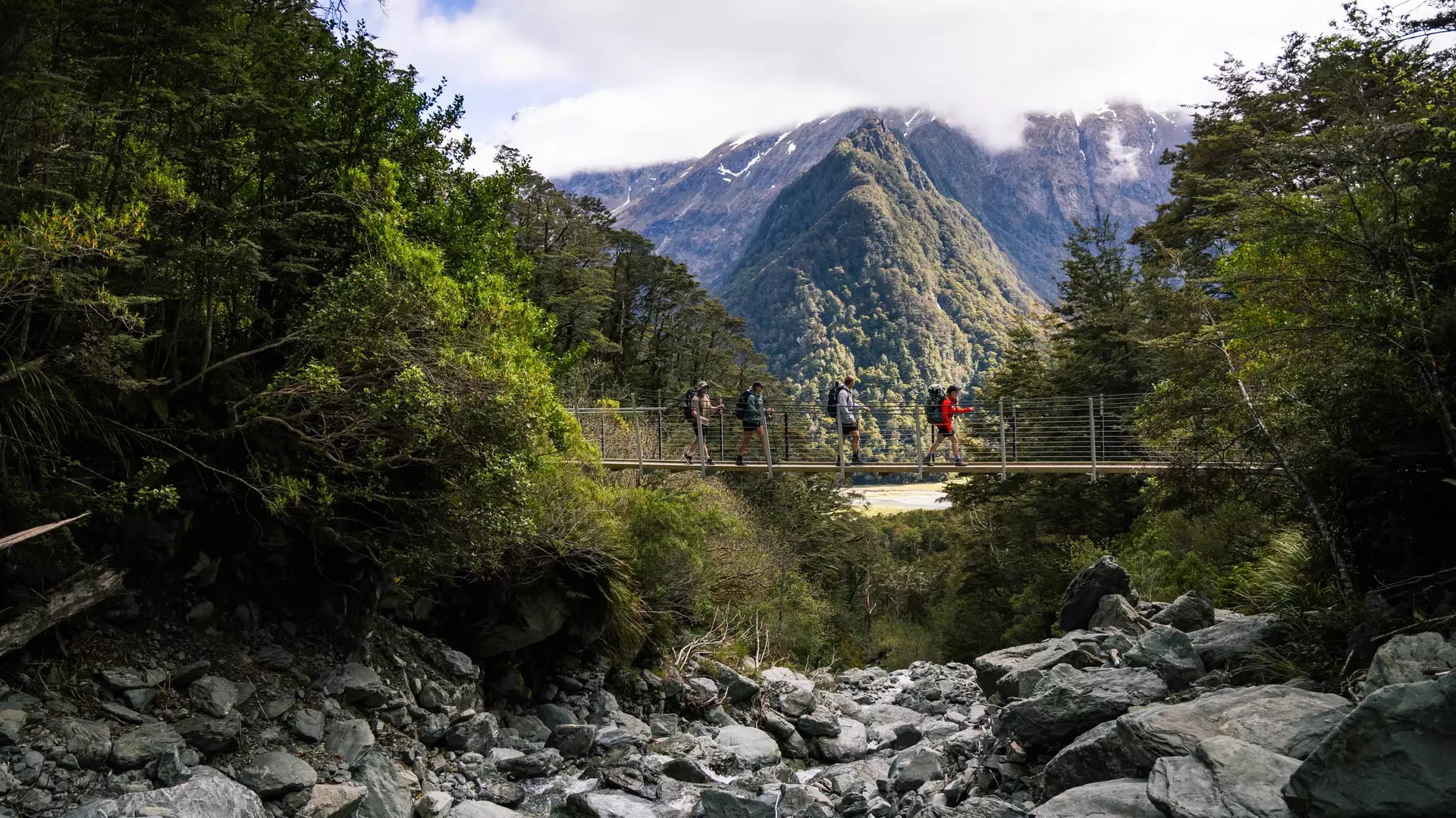 Four hikers crossing a suspension bridge over rocky riverbed with mountains and forest behind