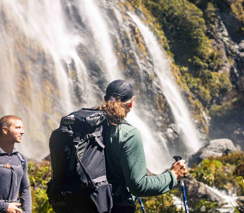 Hikers with backpacks and walking poles stand near a large waterfall in a forested area.