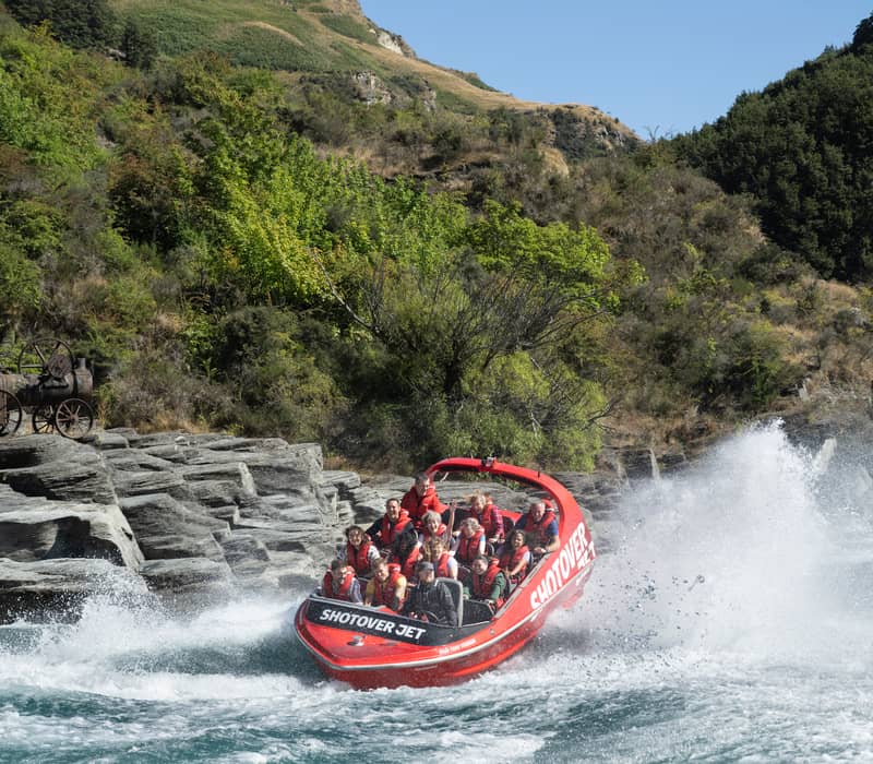 Red Shotover Jet boat speeding on river with passengers wearing life jackets amidst rocky shore and trees.