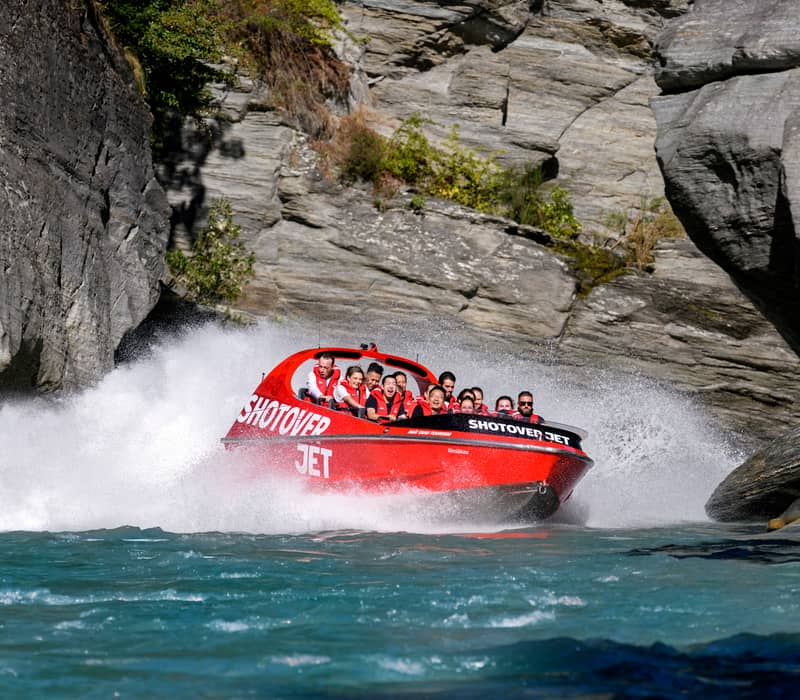 Red Shotover Jet boat speeding through rocky canyon with tourists wearing life jackets and water splashing.