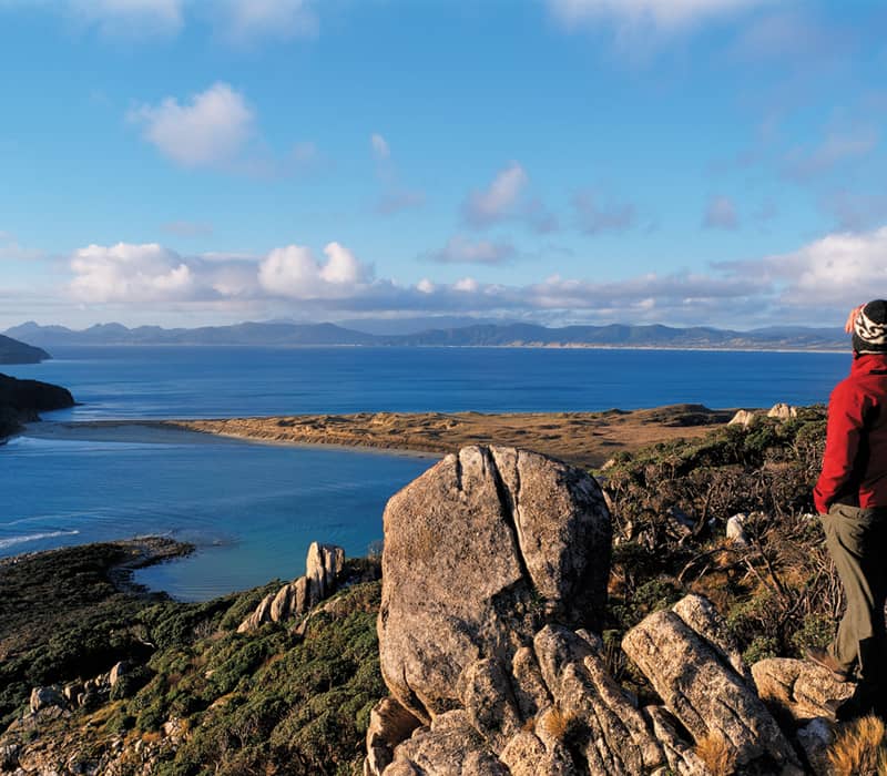 Person in red jacket standing on rocky hill overlooking coastal ocean and hills under a blue sky.