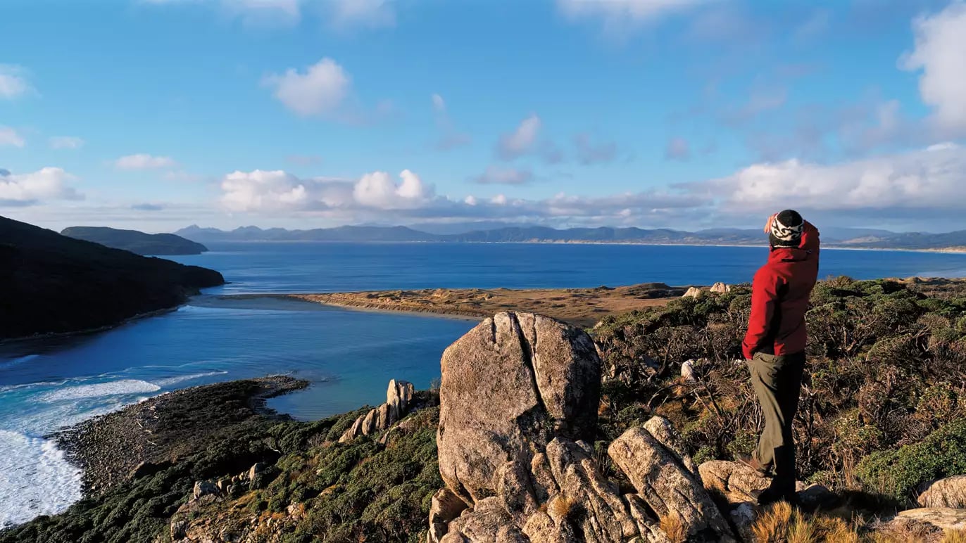 Person in red jacket standing on rocky hill overlooking coastal ocean and hills under a blue sky.