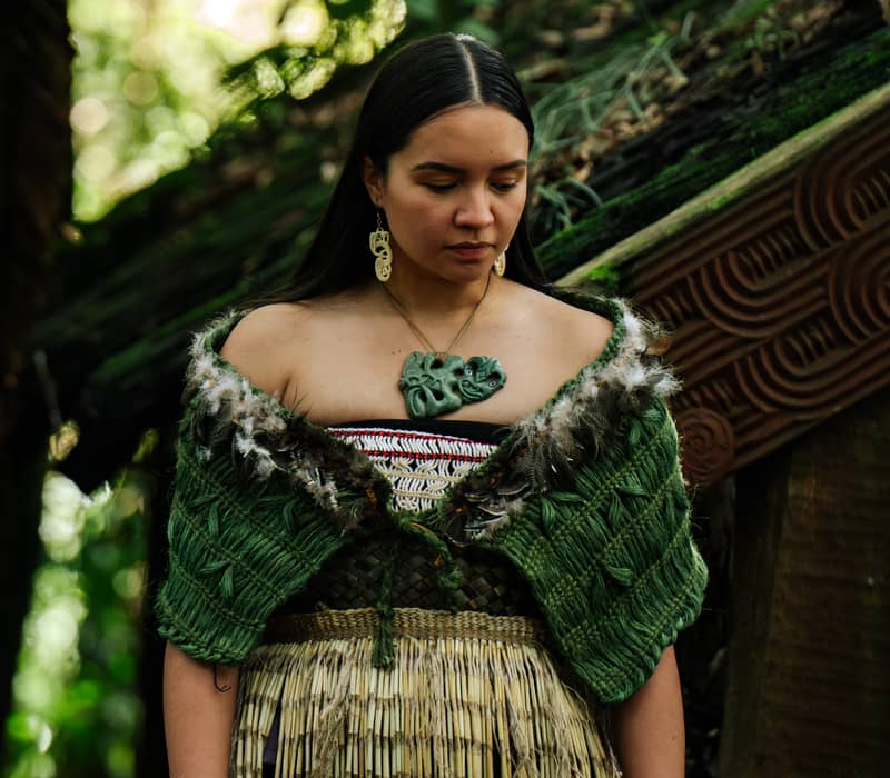 Woman in traditional Maori clothing with green shawl and pendant necklace stands near carved wooden structures.