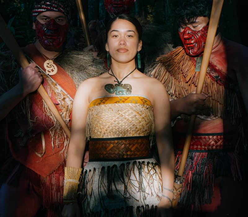 Maori group in traditional dress with face paint and wooden staffs standing in a forest.