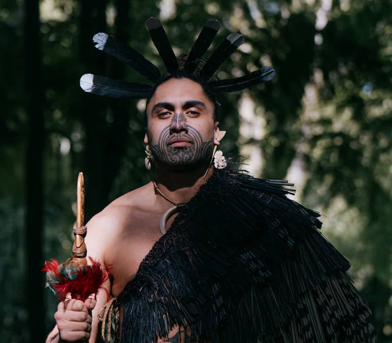 Maori warrior with tattoos and traditional attire holding a spear in a forest setting.