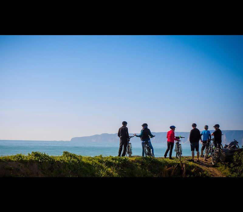 Group of cyclists with helmets standing by the ocean on a grassy cliff under a blue sky.