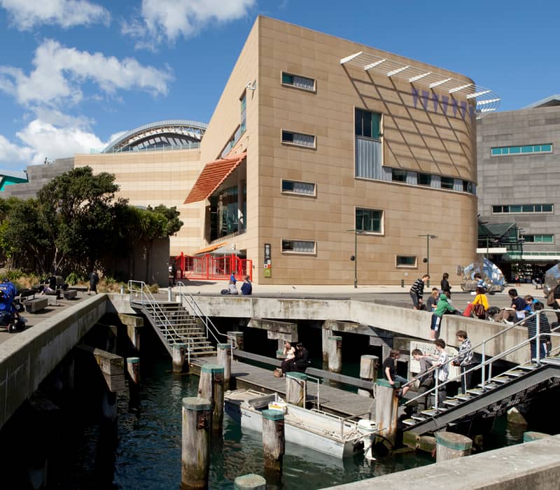 People sitting and standing by the dock and benches outside the modern Te Papa Museum by the water.