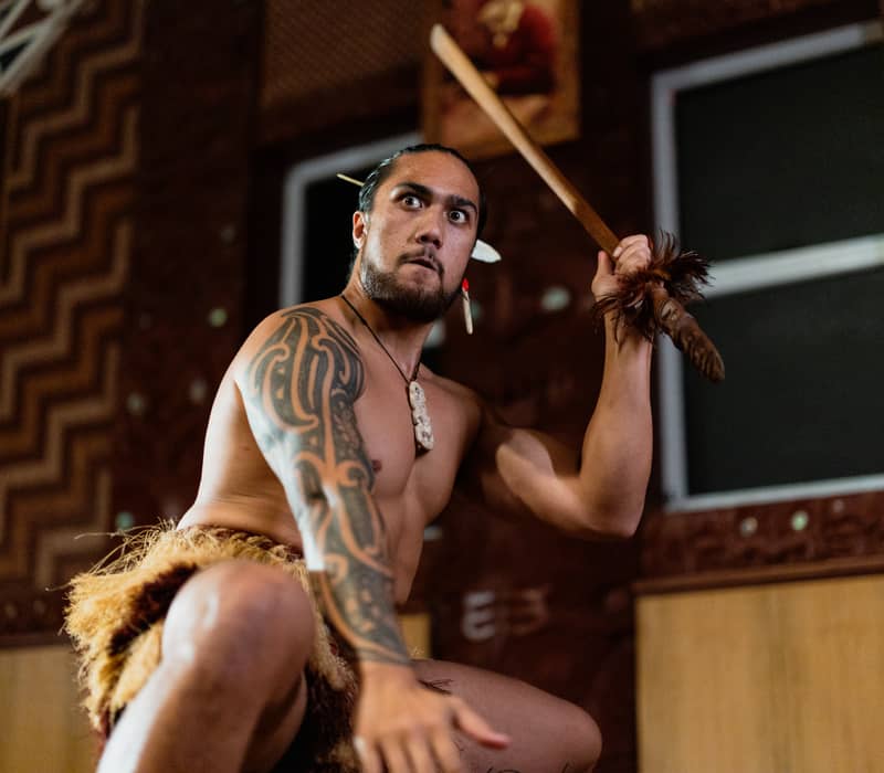 Maori man with tattoos holding spear performing traditional dance indoors.