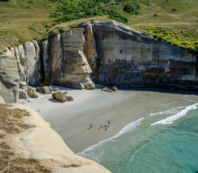 Beach with sandstone cliffs, clear water, and people walking on sand at Tunnel Beach near Dunedin.