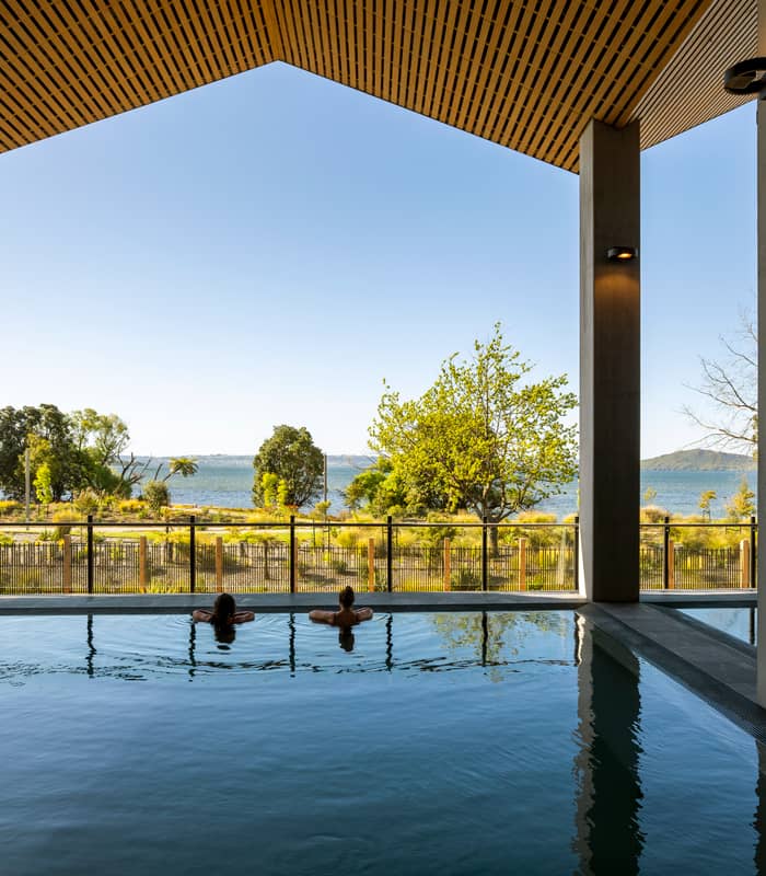 Two people relaxing in an indoor pool with a clear view of a lake and trees outside.