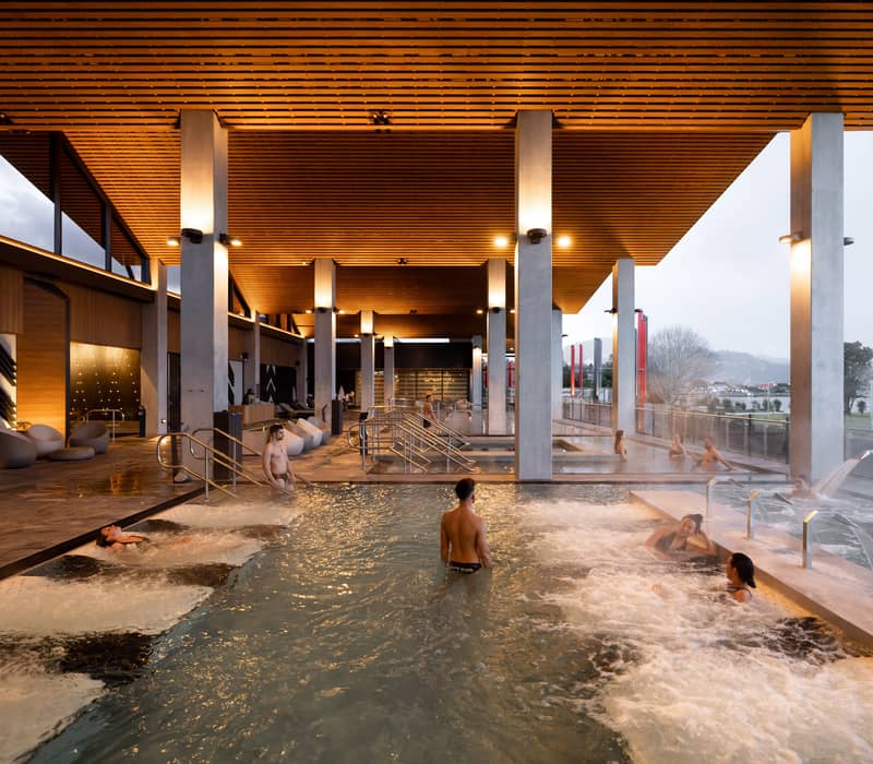 People enjoying hydrotherapy and massage jets in a modern indoor thermal spa pool under a wooden ceiling.