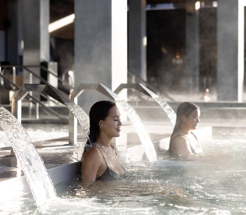 Two women sit under hydrotherapy water jets in a steamy indoor spa pool.