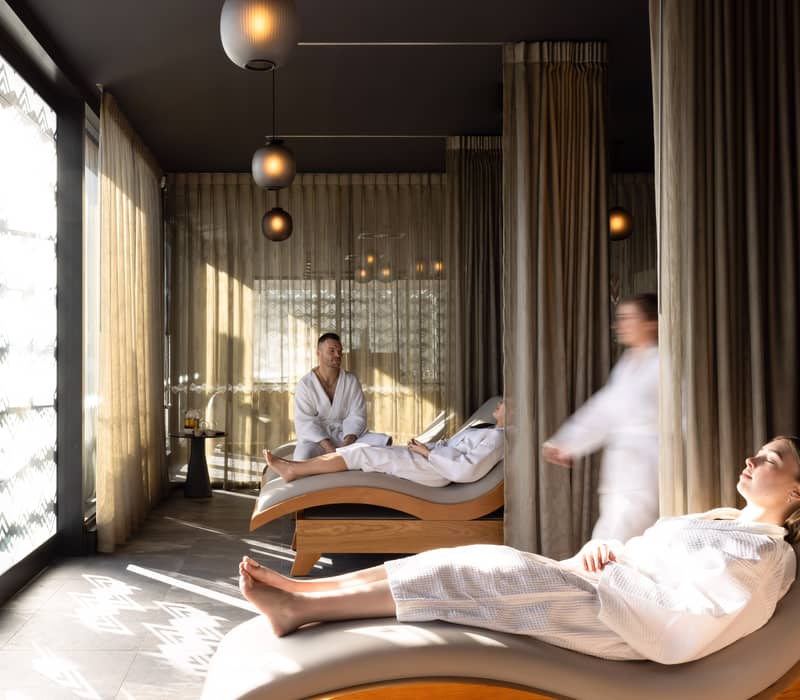 People relaxing on loungers in a spa relaxation lounge with soft natural light and curtains.