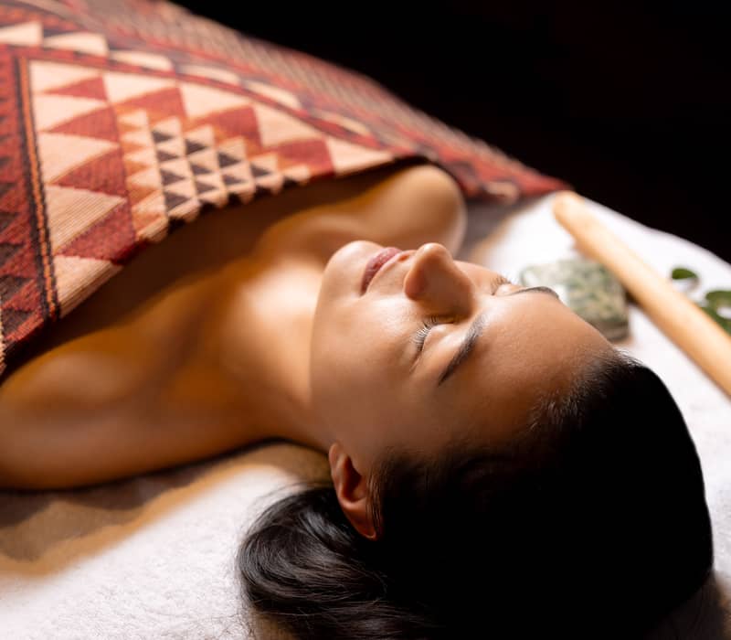 A woman receiving traditional Maori treatment covered with a patterned blanket lying peacefully with eyes closed.