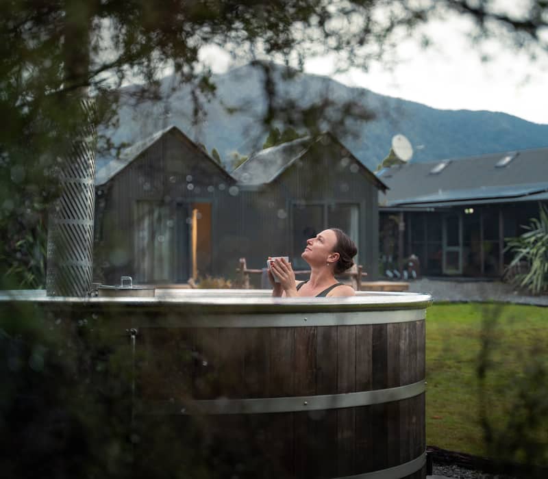 Woman relaxing in outdoor wooden hot tub holding a cup with lodge and mountains in the background