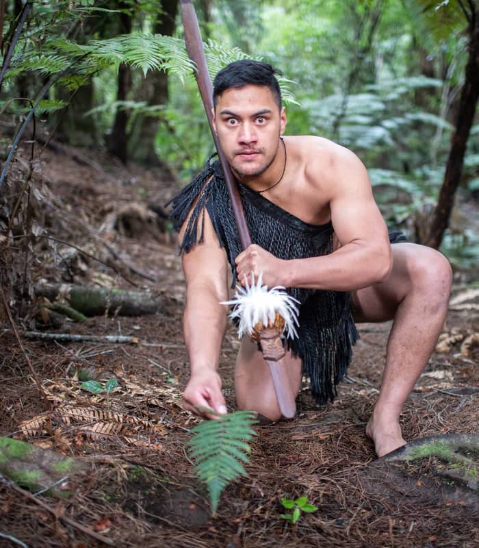 Maori man in traditional attire crouched in forest holding a spear with white feathers.