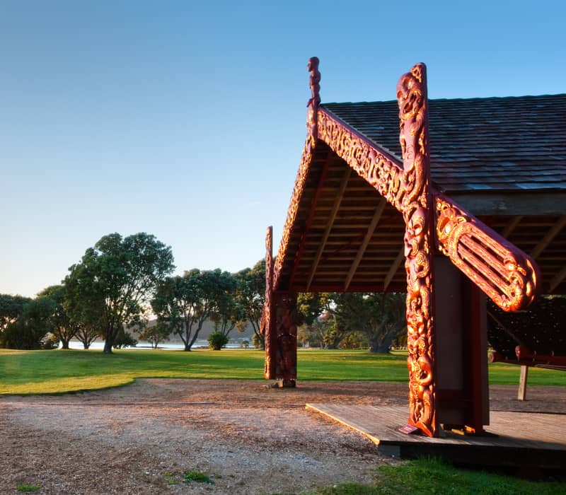 Sunset lighting up a carved Maori shelter near water and trees on a clear day.