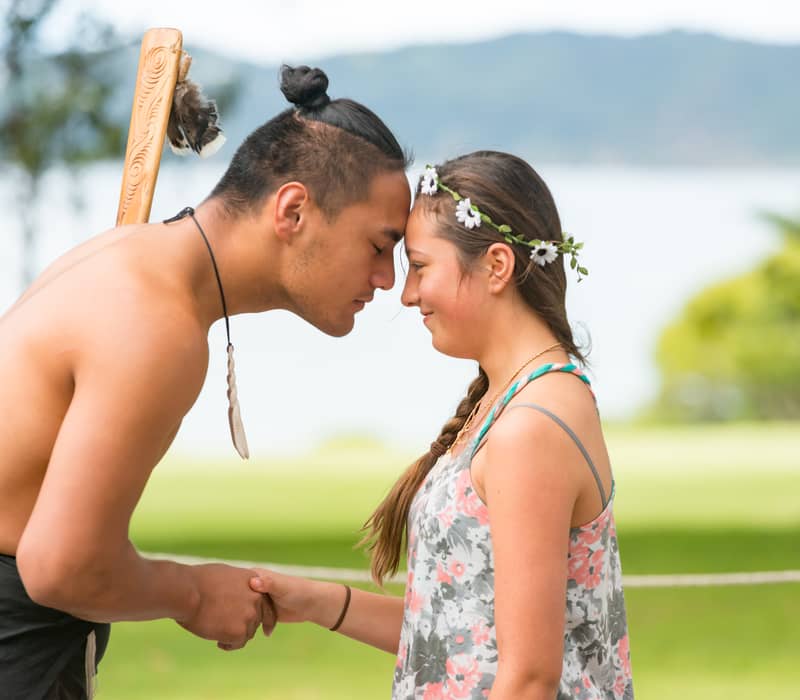 A young man and woman touching foreheads and holding hands in a traditional Maori greeting outside.