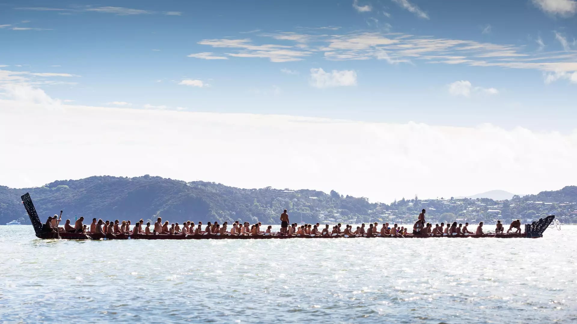 People paddling a long Maori waka canoe on a lake with hills under a blue sky.