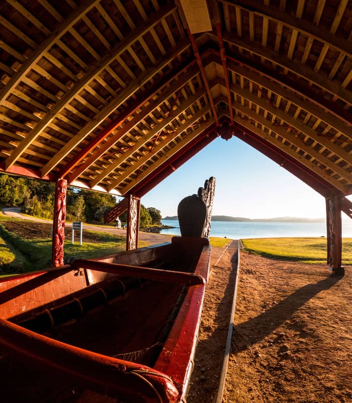 Traditional carved waka under a wooden shelter by a lake at sunset with grassy area and trees.