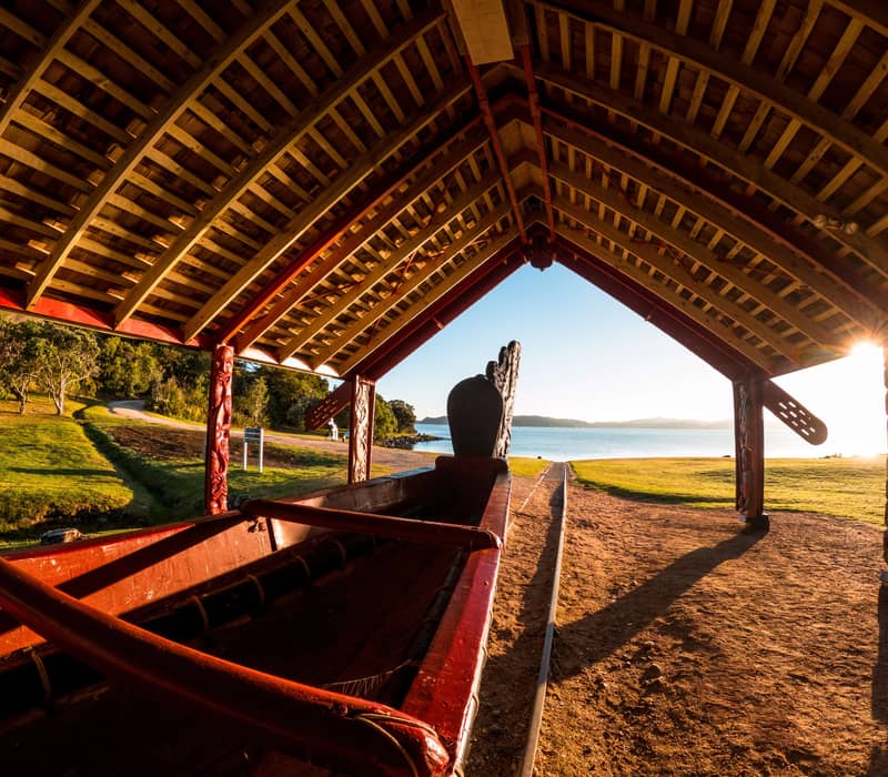 Traditional carved waka under a wooden shelter by a lake at sunset with grassy area and trees.