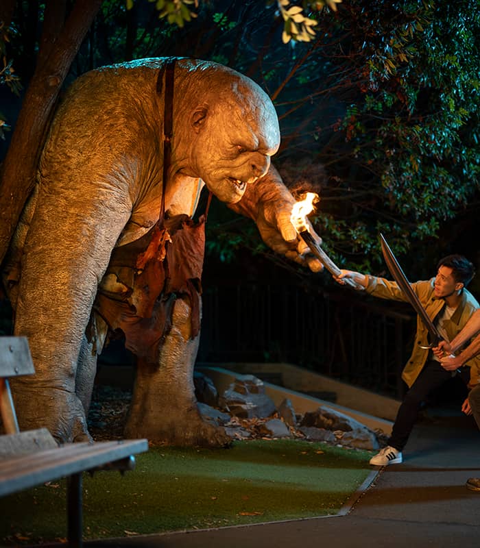 Two men confronting a large troll sculpture holding a torch at night in a park setting.