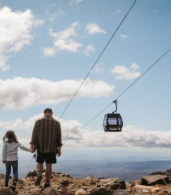Adult and child holding hands hiking on rocky mountain trail near cable car under blue sky with clouds.