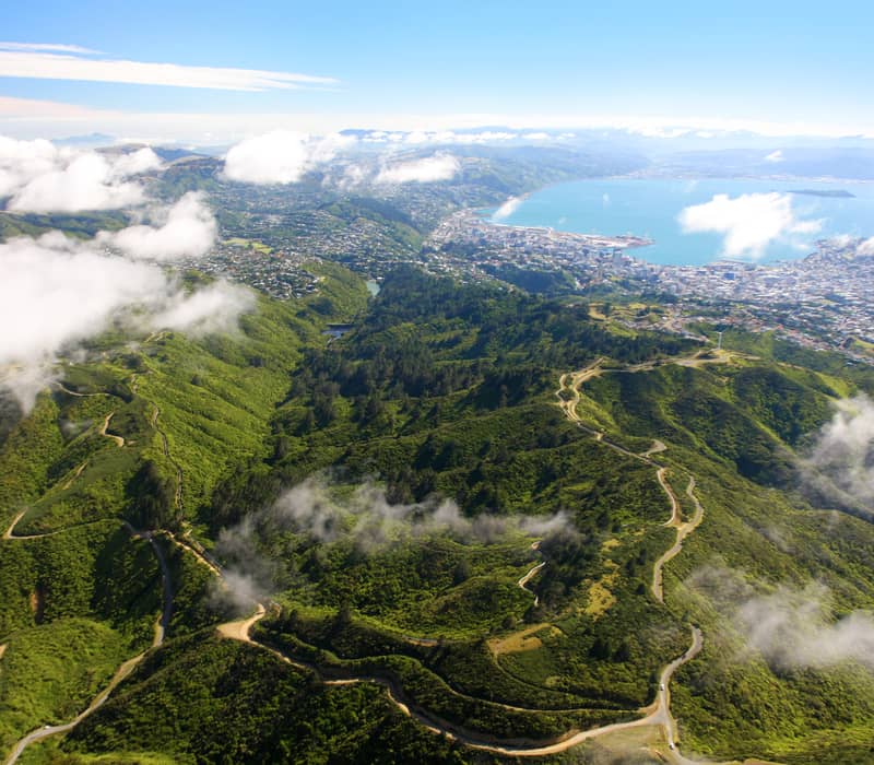 Aerial view of green hills, winding roads, and a coastal city with clouds scattered above.